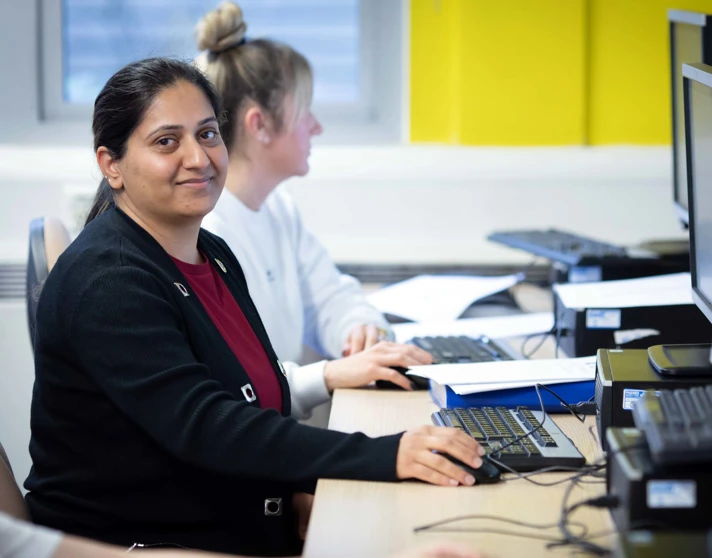 Student in a black cardigan sitting at a computer workstation, smiling while working on a task in a business and admin class. Student in a black cardigan sitting at a computer workstation, smiling while working on a task in a business and admin class.